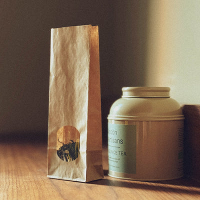 A narrow brown paper bag with a round sticker and a round, light green metal tin with a label, standing on a wooden surface in front of a dark background.