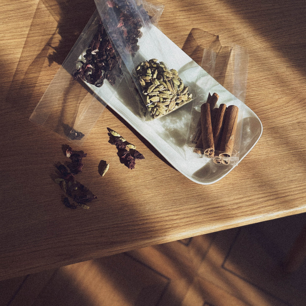 Top view of an oblong white bowl standing on a light wooden surface and containing various spices in transparent plastic bags: dried hibiscus flowers, green cardamom pods, and cinnamon sticks.